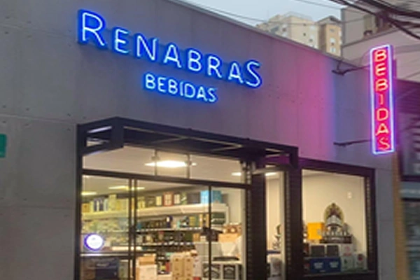 Fachada da loja Renabras Bebidas, com letreiros em neon azul e rosa iluminando a entrada.