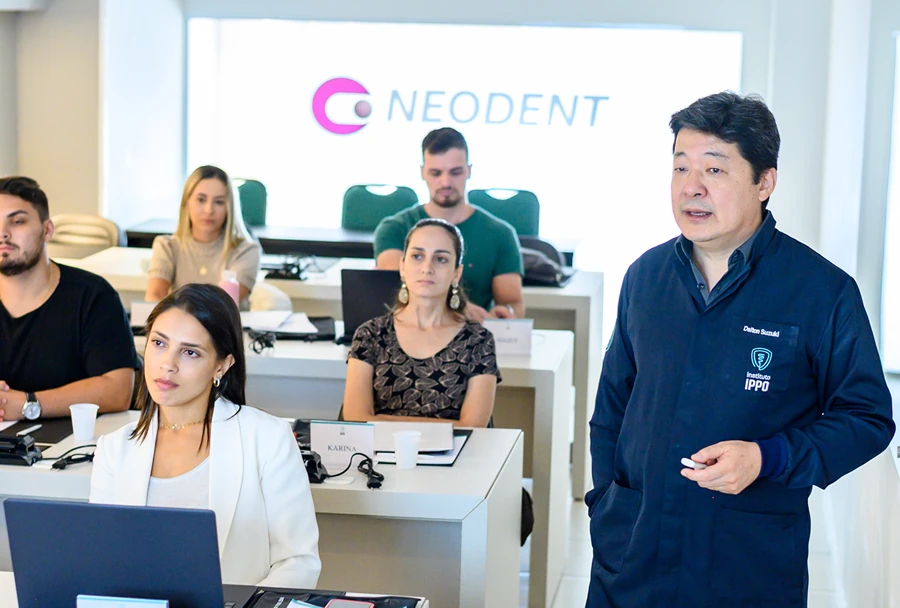 Instrutor do Instituto IPPO, de jaleco azul, conduzindo uma aula enquanto os alunos ouvem atentamente, com o logotipo da Neodent ao fundo.