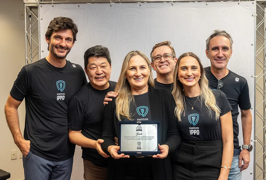 Grupo de seis sorridentes do IPPO, em camisetas pretas com logo, mulher no centro segura placa dos 10 anos, fundo branco e metálico.