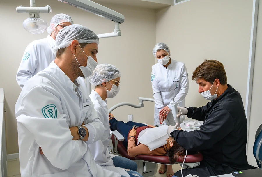 Equipe de dentistas ao redor de uma cadeira odontológica observando um procedimento, com o paciente deitado e o dentista realizando o tratamento.