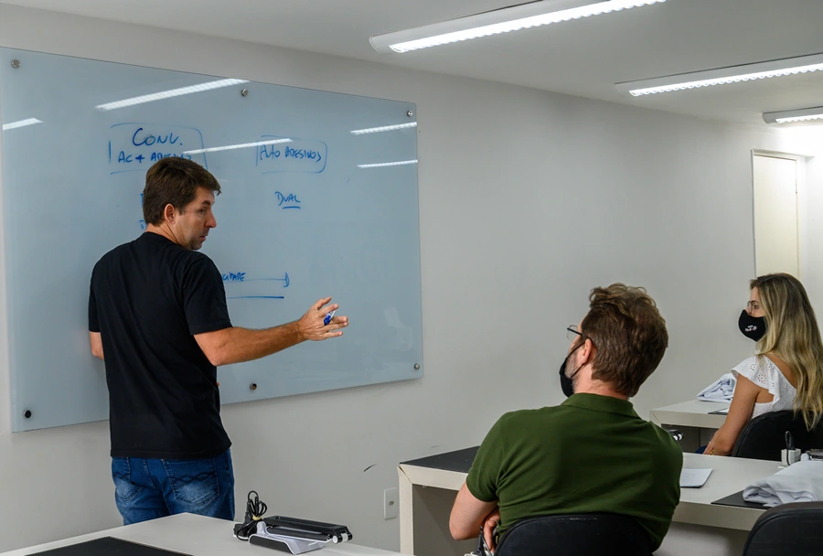 Professor escrevendo no quadro branco enquanto alunos observam, em sala de aula com ambiente iluminado e ar-condicionado no teto.