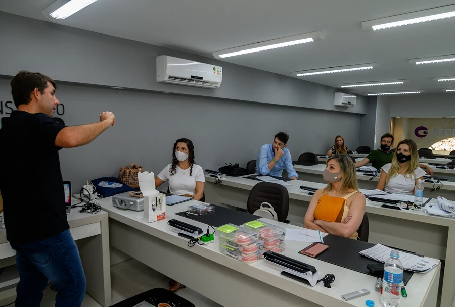 Professor gesticulando com as mãos para alunos que prestam atenção, em sala de aula com ar-condicionado e ambiente claro.