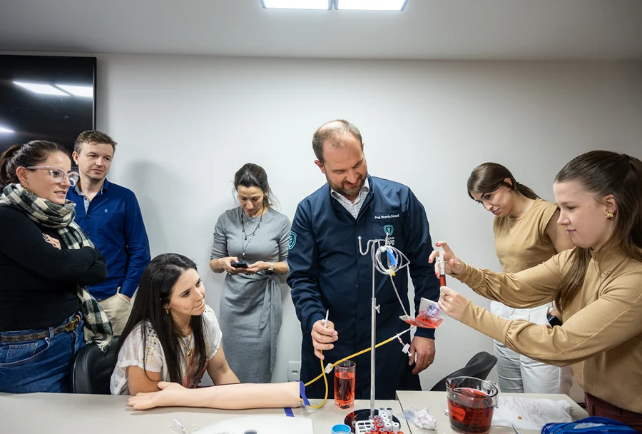 Professor Ricardo Demétrio realizando uma demonstração de transfusão simulada, com alunos observando atentamente o procedimento e participando.